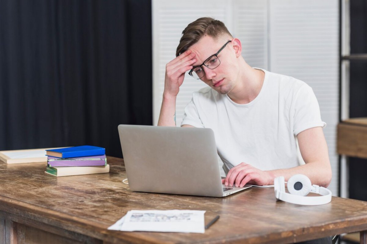 A person sits at a wooden table, looking stressed while working on a laptop, with books and headphones nearby.