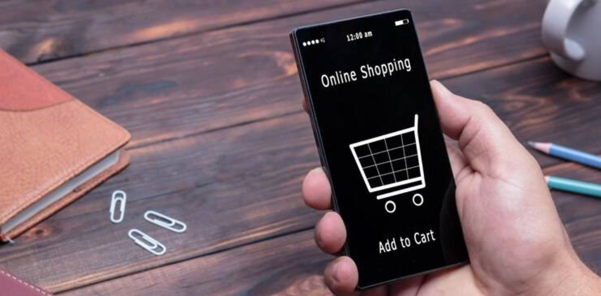 A hand holds a smartphone displaying Online Shopping with a shopping cart icon, surrounded by office supplies on a wooden desk.