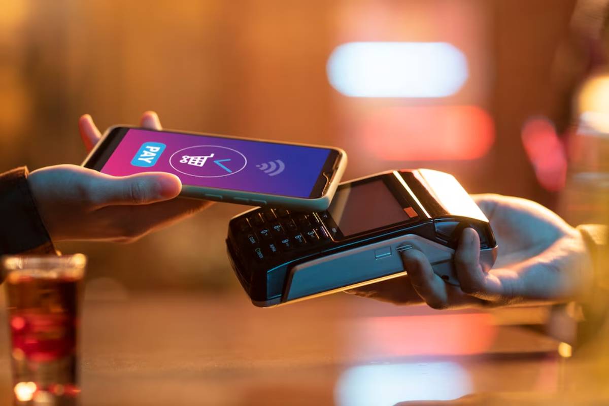 A hand holds a smartphone displaying a payment screen, hovering over a card reader at a bar, with a drink visible in the foreground.