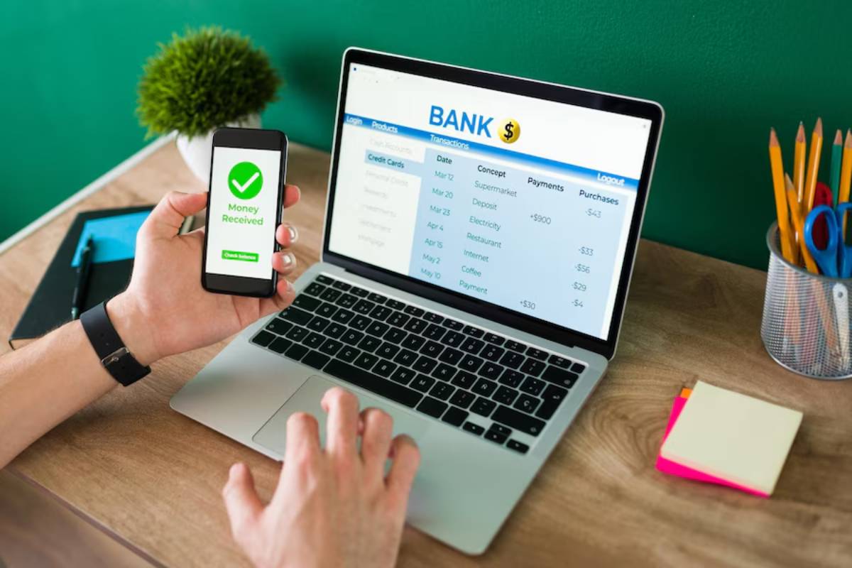 A person holds a smartphone displaying Money Received while viewing bank transactions on a laptop, with office supplies nearby.