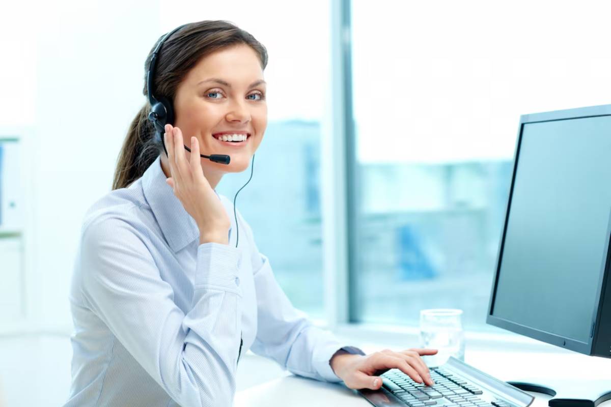 A woman wearing a headset sits at a desk with a computer, focused on her work in a bright, modern office environment.