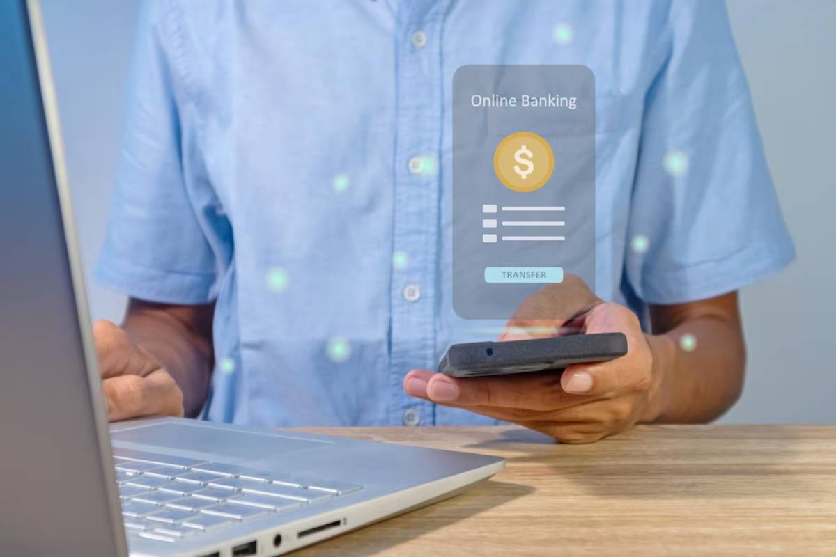 A person in a blue shirt uses a smartphone to perform online banking transactions, with a laptop open in front of them.