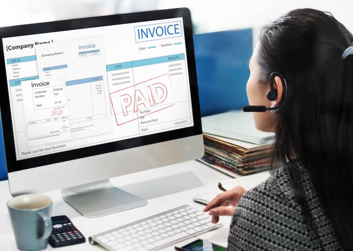A woman with a headset reviews a paid invoice on a computer screen, surrounded by office supplies and paperwork.