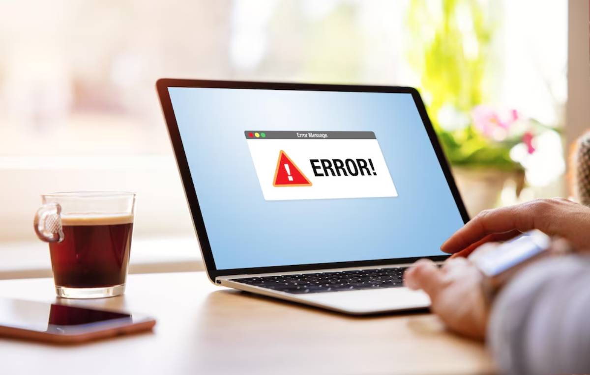 A person holding a phone beside a laptop displaying an error message on the screen, with a cup of coffee nearby.