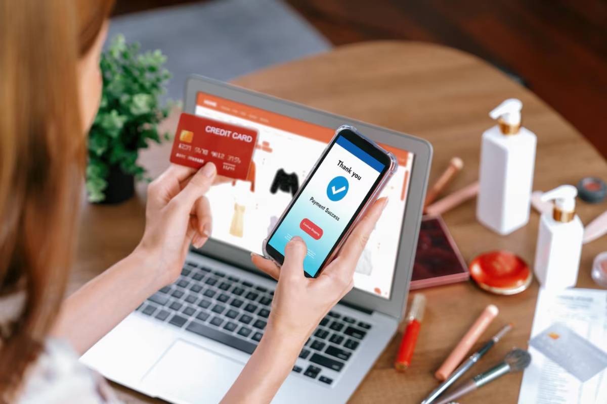 A person holds a smartphone displaying a Payment Success message, with a credit card in hand and a laptop showing an online shop.