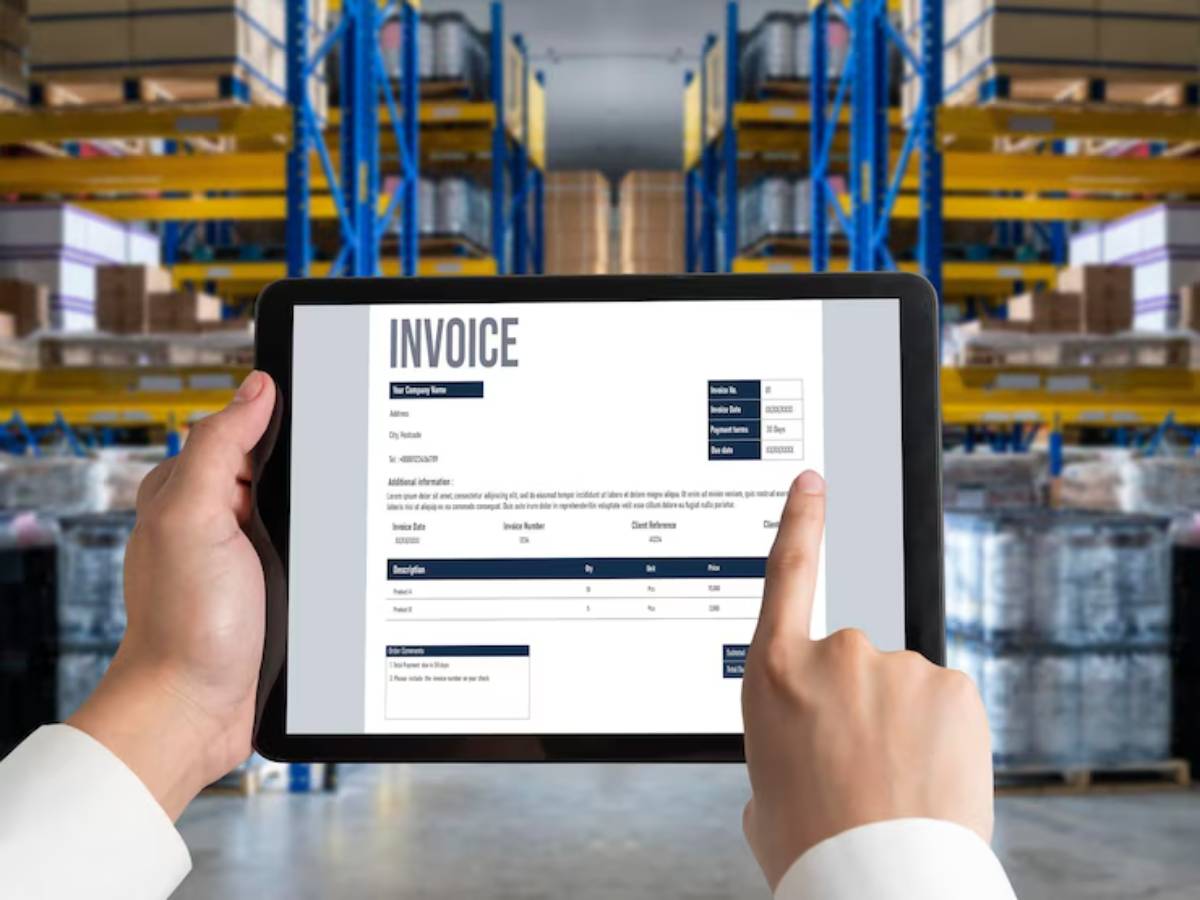 A person holding a tablet displaying an invoice, with a warehouse backdrop filled with storage shelves and packaging.