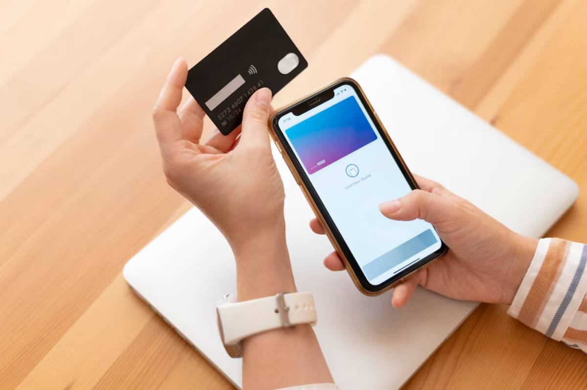 A person holds a smartphone displaying a digital payment app while using a contactless credit card on a wooden table.