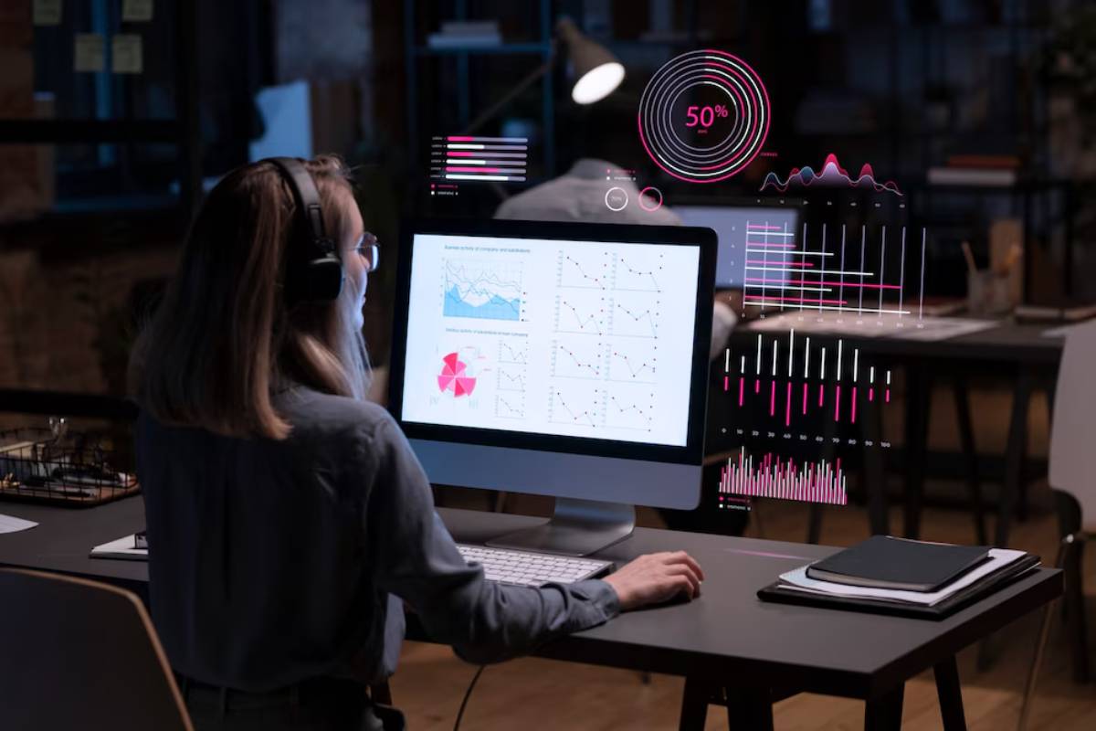 A woman wearing headphones analyzes data visualizations and charts displayed on her computer screen in a dimly lit office.