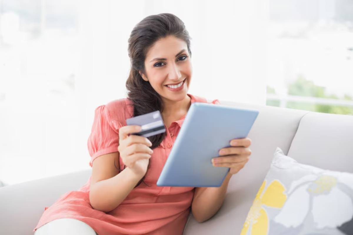 A woman in a pink top holds a credit card and a tablet, sitting on a couch with a floral pillow, in a bright room.