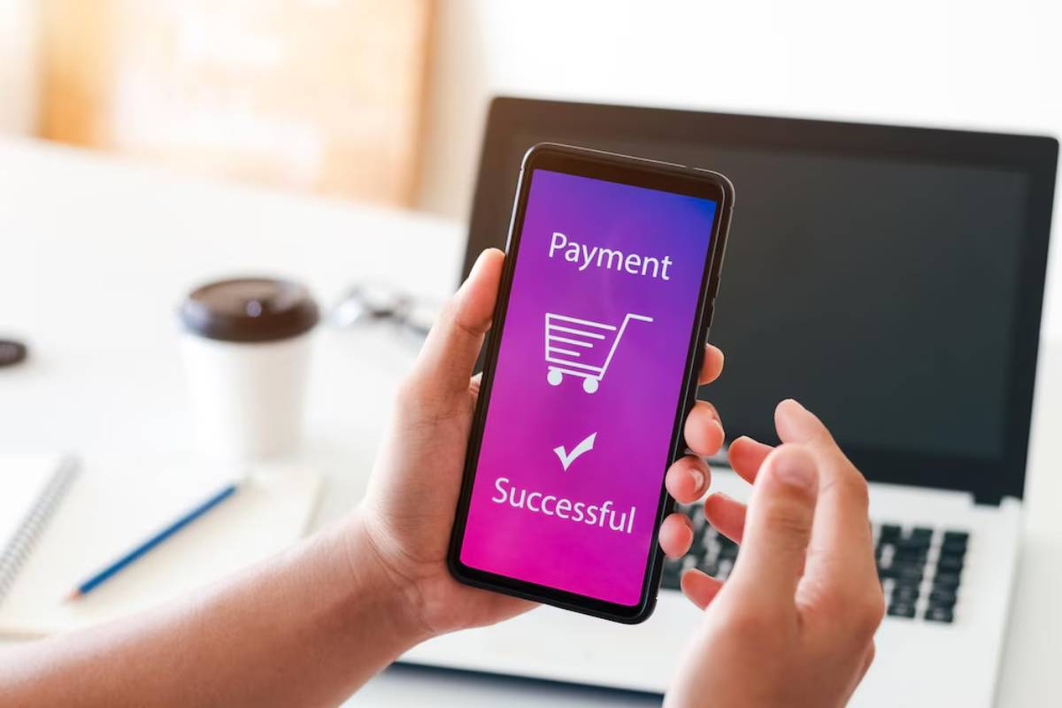 A person holds a smartphone displaying a successful payment confirmation, with a shopping cart icon and checkmark, beside a laptop.