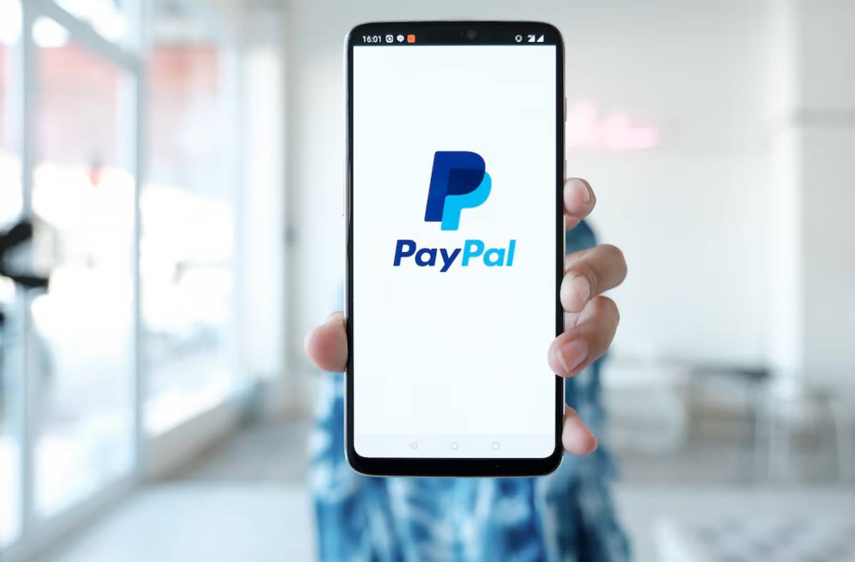 A person holds a smartphone displaying the PayPal logo in a bright, modern environment.