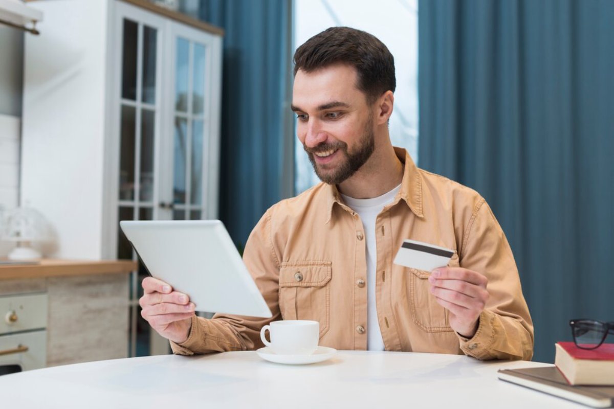 A person in a beige jacket holds a tablet in one hand and a credit card in the other, seated at a table with a coffee cup.