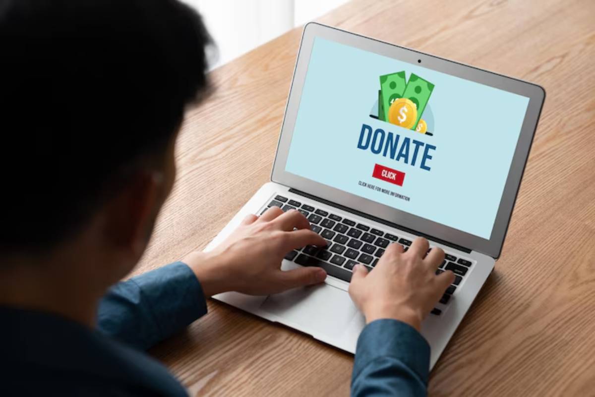 A person in a blue shirt is using a laptop with a DONATE button and dollar bills graphic on the screen, encouraging online donations.