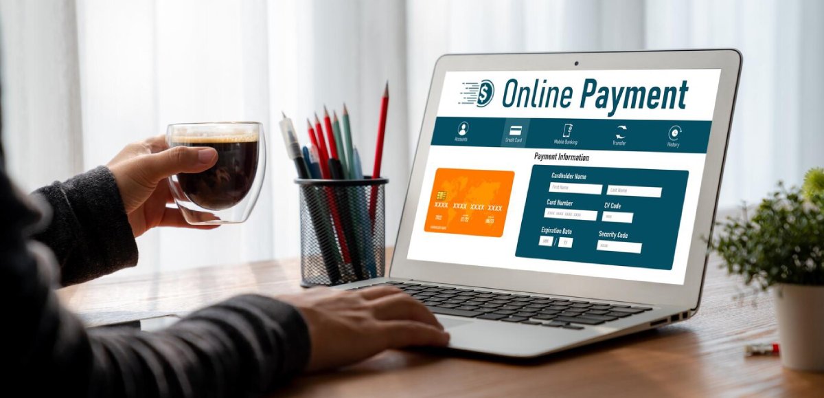 A person sips coffee while using a laptop displaying an online payment interface with credit card and security fields.