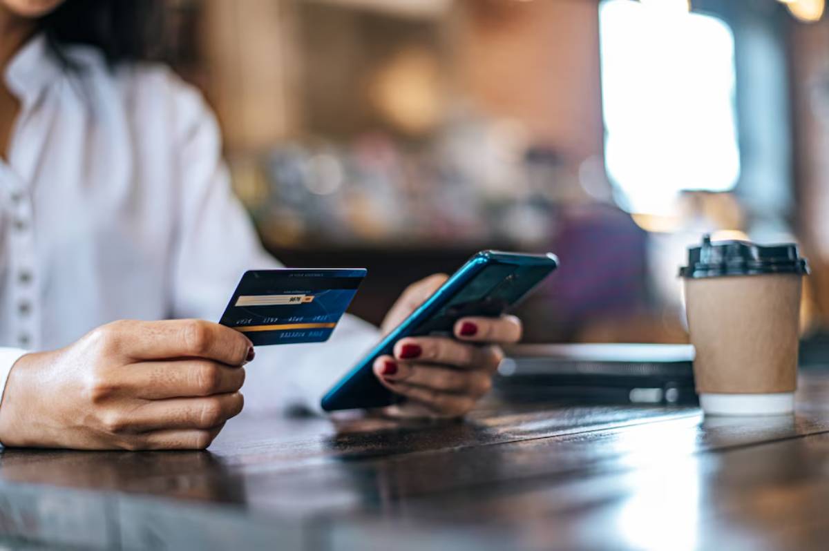 A woman holding a credit card in one hand and a smartphone in the other, sitting at a café table with a coffee cup nearby.