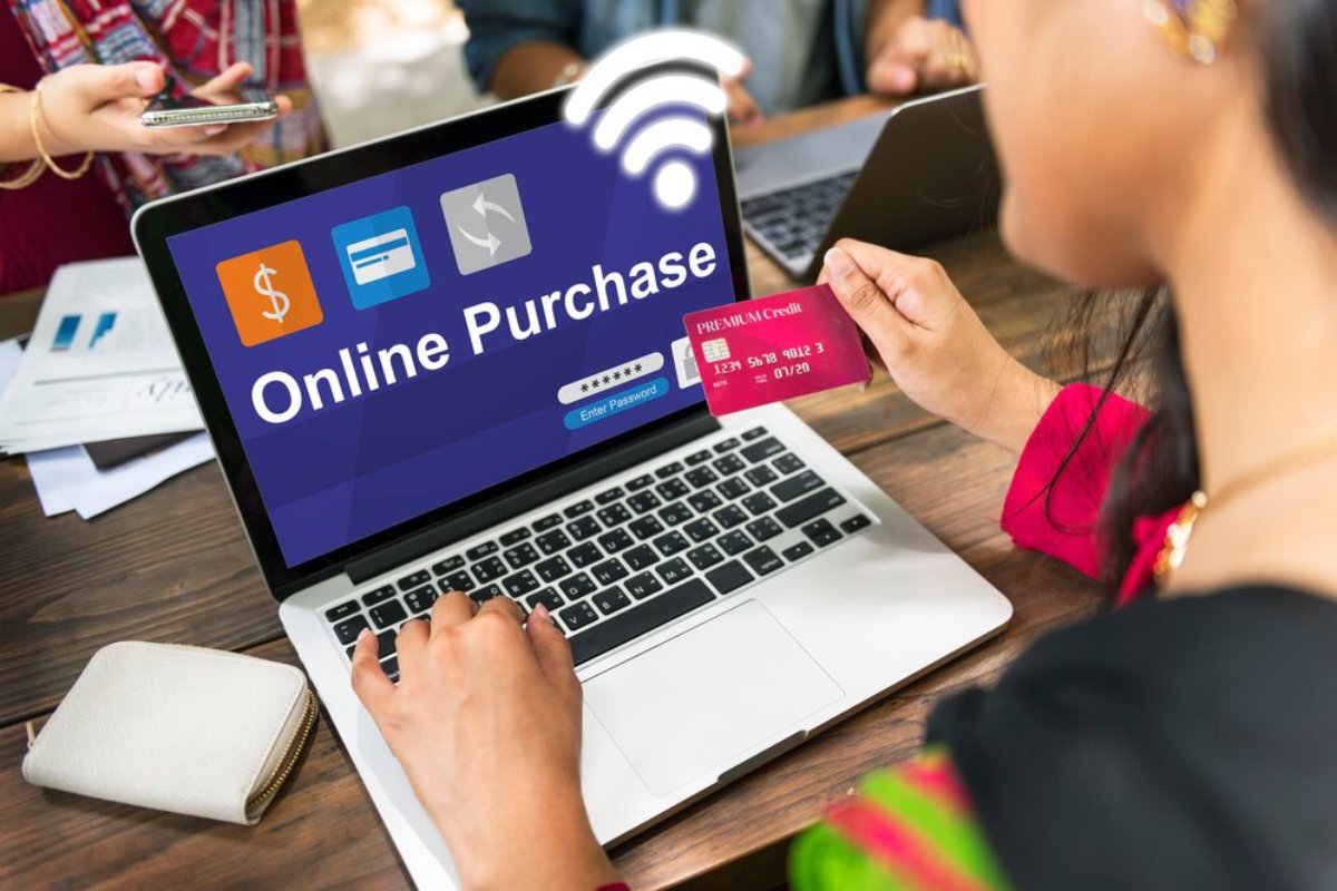 A person uses a laptop to make an online purchase, holding a credit card, while another individual is seen in the background.