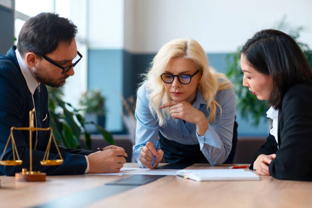 Three professionals are engaged in a discussion over documents on a conference table, with a scale in the foreground.