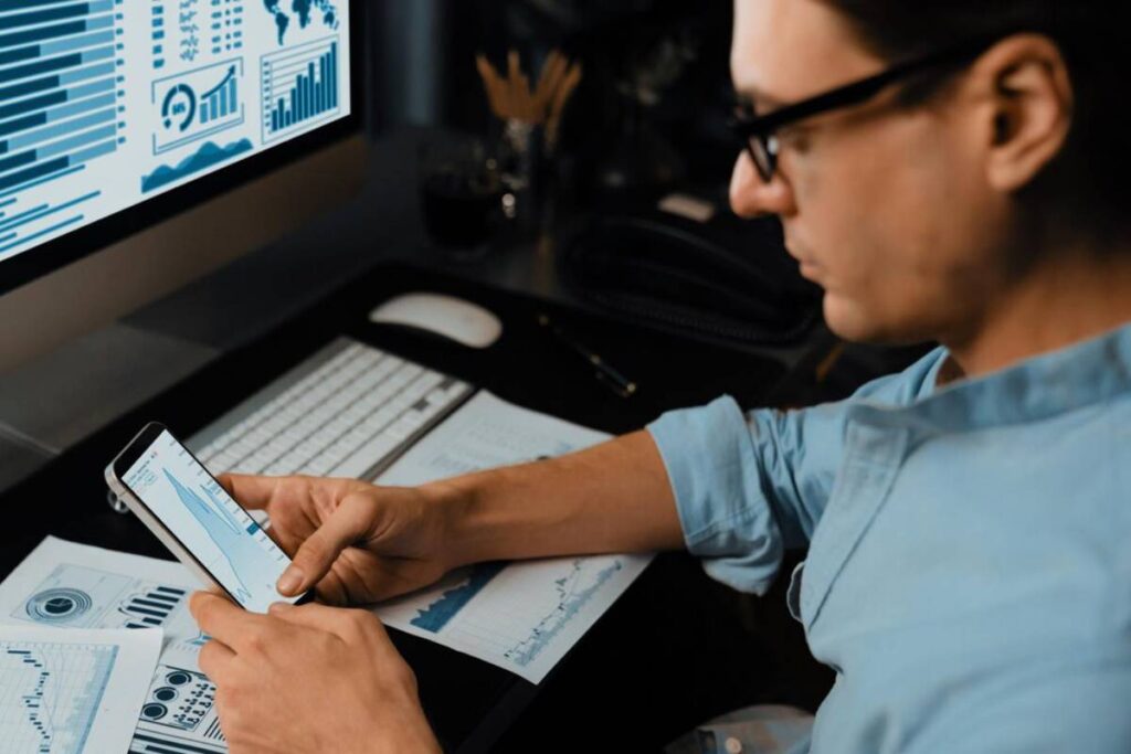 A person analyzes data on a smartphone while sitting at a desk with graphs and charts printed on papers and a computer monitor displaying data.