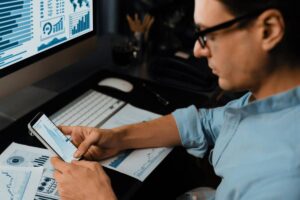 A person analyzes data on a smartphone while sitting at a desk with graphs and charts printed on papers and a computer monitor displaying data.