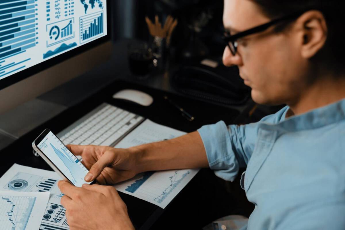 A person analyzes data on a smartphone while sitting at a desk with graphs and charts printed on papers and a computer monitor displaying data.