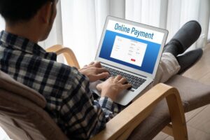 A person in a plaid shirt sitting comfortably, using a laptop to complete an online payment, with a bright window in the background.