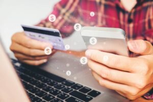 A person holding a smartphone and a credit card, with dollar symbols floating around, while using a laptop for online shopping.