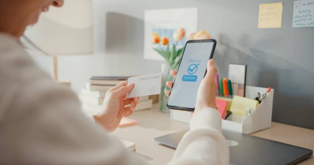 A person holds a credit card and a phone displaying a successful payment message, surrounded by office supplies and flowers.