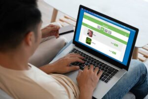 A person is using a laptop to shop online for groceries, displaying items like meat and lettuce with price tags.