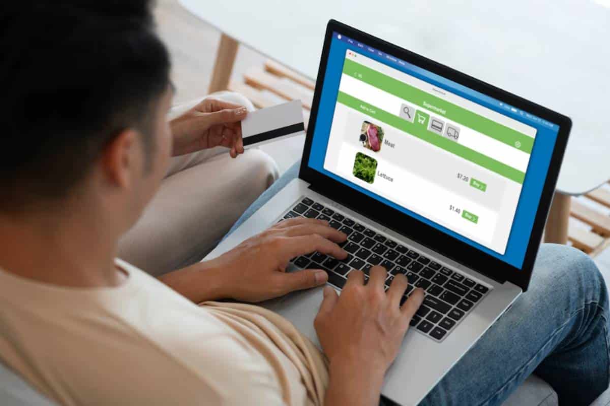A person is using a laptop to shop online for groceries, displaying items like meat and lettuce with price tags.
