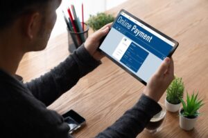 A person holds a tablet displaying an online payment interface, with a coffee cup and small plants on a wooden table.