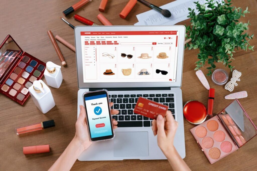 A person shopping online with a laptop and smartphone, surrounded by makeup products and a credit card, celebrating a successful payment.