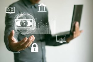 A man in a gray shirt holds a laptop and gestures towards floating financial icons, including money, banking, and data security symbols.